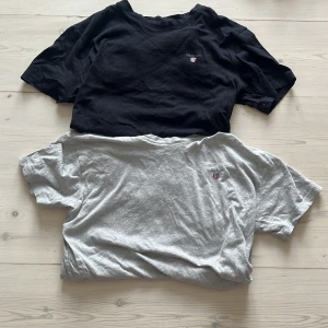 2-pack GANT t-shirts svart & grå - Två klassiska t-shirts från GANT i storlek 158/164. En svart och en ljusgrå, båda med rund hals och diskret GANT-logo broderad på bröstet. Sköna och stilrena, perfekta till jeans eller shorts. Tillverkade i mjuk bomull för maximal komfort.