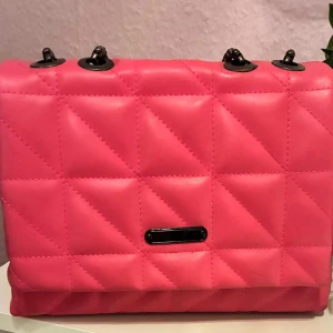 Quiltad rosa axelväska med kedja - Säljer en snygg quiltad axelväska i starkt rosa med svarta metalldetaljer och kedjerem. Väskan har en rektangulär form och är tillverkad i syntetmaterial. Perfekt för dig som vill sticka ut med en färgglad accessoar.