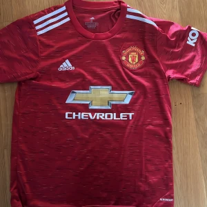 Manchester United Adidas matchtröja S - Röd Manchester United fotbollströja från Adidas med vita detaljer och klubbmärke på bröstet. Tröjan har korta ärmar, klassiska Adidas-ränder på axlarna och Chevrolet-logga framtill. Tillverkad i lätt och ventilerande AEROREADY-material.