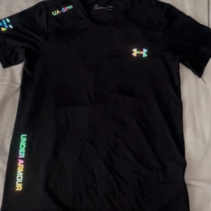 Svart Under Armour t-shirt M - Svart t-shirt från Under Armour med färgglada loggor och textdetaljer. Modellen är kortärmad och har en normal passform. Tillverkad i ett lätt och snabbtorkande syntetmaterial, perfekt för träning och aktiva dagar.