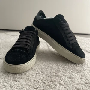 Svarta Axel arigato skor  - Stilrena svarta sneakers i mocka med klassisk vit platt sula. Skorna har rund tå, svarta skosnören och detaljer i svart skinn vid hälen. Perfekta för dig som gillar en clean och enkel look med lite lyxigare känsla. Priset kan även diskuteras 😁😁