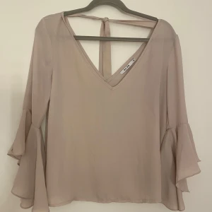 Beige blus med volangärm från NA-KD - Supersnygg beige blus från NA-KD med v-ringning och volangärmar. Blusen har knytband i nacken.
