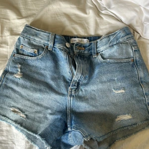 Blå jeansshorts från Zara - Snygga blå jeansshorts från Zara med slitna detaljer och råa kanter. Klassisk femficksmodell med dragkedja och knapp framtill. Perfekta för varma dagar och ger en avslappnad vibe.