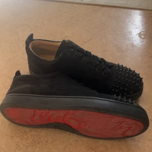 Christian Louboutin svarta sneakers med nitar - Svarta sneakers från Christian Louboutin i mocka med ikoniska röda sulor och svarta nitar över tån. Skorna har snörning och en låg siluett, perfekt för dig som vill sticka ut med en edgy vibe. Snygg kontrast mellan svart ovandel och röd undersida. Några nitar borta men kan köpa billiga på nätet. För små för mig 