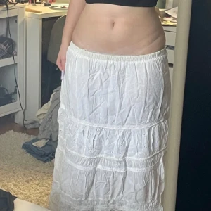 Vit långkjol med broderade detaljer - En vit långkjol i bomull med broderade detaljer och volanger. Kjolen har resår i midjan och flera lager med fina spetsinslag. Perfekt för dig som gillar en romantisk stil!