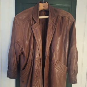 Brun skinnjacka från Soki Leather - Säljer en brun lång skinnjacka från Soki Leather med bred krage och stora fickor. Jackan har en klassisk, avslappnad passform och är tillverkad i äkta skinn. Perfekt för dig som vill ha en tidlös och cool look med vintagekänsla.