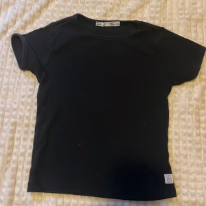 Svart t-shirt från lager 157 - En stilren svart t-shirt från Ettfemsju med klassisk passform och korta ärmar. T-shirten har en enkel design med rund halsringning och en liten vit logolapp nertill. Perfekt till jeans eller shorts för en clean look.