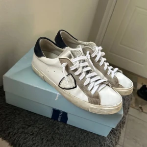 Philippe Model vita och beige sneakers - Snygga sneakers från Philippe Model i vitt skinn med beige mockadetaljer och mörkblå häl. Klassisk låg modell med vit sula och snörning. Ikonisk sköldlogga på sidan och bekväm platt sula. Perfekta för en stilren och avslappnad look.