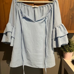 Ljusblå offshoulder blus från Hailys (affär i Grebbestad) - Supersöt ljusblå blus med offshoulder-modell och volangärmar i två lager. Blusen har knytband framtill och en luftig, lätt struktur. Perfekt för dig som gillar romantiska och trendiga detaljer.