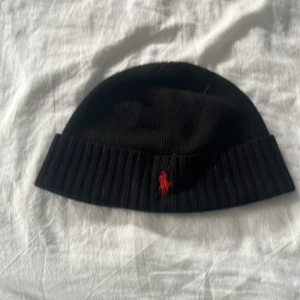Svart merinoullsmössa Polo Ralph Lauren - Pris kan diskuteras också. Svart ribbstickad mössa från Polo Ralph Lauren i mjuk och tvättbar merinoull. Klassisk design med uppvikt kant och röd broderad logga framtill. Perfekt accessoar för kalla dagar och snygg till streetwear.