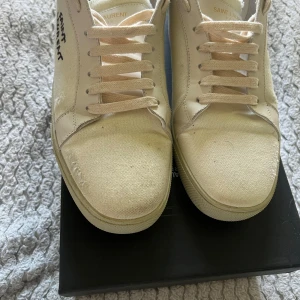Saint Laurent vita sneakers i canvas - Saint Laurent sneakers i vit canvas med snörning och diskret logga på sidan. Klassisk låg modell med rund tå och platt sula. Ovandel i canvas och detaljer i vitt skinn. Perfekta för en clean och stilren look.