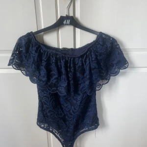 Mörkblå offshoulder spetsbody - Säljer en mörkblå offshoulder body från Only One i vacker spets med blommigt mönster. Toppen har bred volang runt axlarna och är helt ärmlös, perfekt för en trendig look. Sitter tight och har en feminin, romantisk vibe.