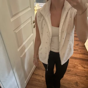 Vit fleeceväst med pärldetaljer - Säljer en vit fleeceväst med hög krage och dragkedja framtill. Västen är dekorerad med små pärlor över hela framsidan. Storlek M men passar bra med S 