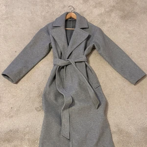 Grå kappa med bälte och stora slag - Snygg grå kappa med breda slag och knytskärp i midjan. Lång modell med raka ärmar och två sidofickor. 