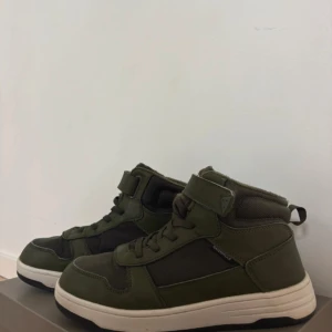 Gröna höga sneakers med kardborre - Snygga höga sneakers i mörkgrönt och svart med vit sula. Skorna har snörning och kardborreband upptill för extra support. Ovandelen är i en mix av syntet och textil, vilket ger en cool kontrast. Perfekta för dig som gillar streetstyle och vill sticka ut.