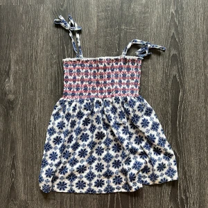 Topp med knytband XS - Supersöt topp med smock upptill och knytbara axelband. Vit botten med blått blommönster och röda detaljer. Perfekt för varma dagar och har en luftig passform. Tillverkad i mjuk bomull som känns skön mot huden.