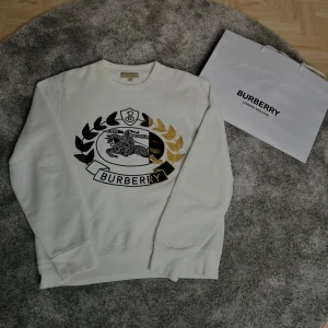 Vit sweatshirt från Burberry M - Vit sweatshirt från Burberry med stort broderat emblem framtill i svart och guld. Klassisk rund hals och långa ärmar. Tröjan är i bomullsmix och har en clean, lyxig vibe med snygga detaljer och logga framtill.