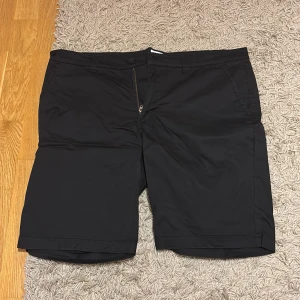 Svarta shorts från FOUR•D, XL - Klassiska svarta shorts från FOUR•D i storlek XL. De har en rak passform, bälteshällor, sidofickor och en bakficka med knapp. Stängs med dragkedja och knapp framtill. Perfekta för varma dagar när du vill ha en clean och enkel look.
