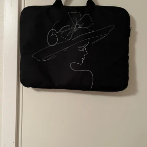 Svart datorväska med konstmotiv - Snygg svart datorväska med ett stilrent vitt linjemotiv av en kvinna med hatt på framsidan. Väskan har handtag upptill och dragkedja längs kanten. Perfekt för att bära din laptop med stil.