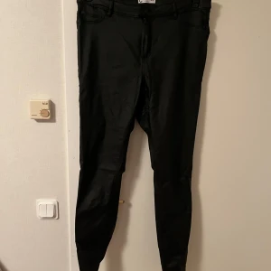 Svarta skinny byxor Jane XL/32 - Svarta skinny byxor från Cubus i modellen Jane. Byxorna har en tight passform och klassisk femficksdesign. Materialet har en lätt glansig finish som ger en cool look. Perfekta för dig som gillar en smal siluett och vill ha ett par stilrena svarta byxor.