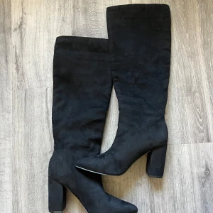 Svarta höga mockastövlar med klack - Snygga svarta stövlar i mocka-look med hög, bred klack och spetsig tå. Stövlarna går upp till knäet och har en stilren, minimalistisk design som passar perfekt till kjol eller jeans. Perfekta för dig som vill ha en trendig och elegant look.