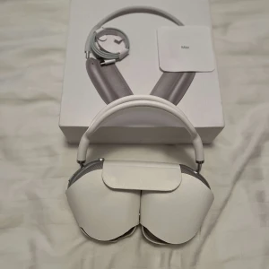 Airpod Max vit - Bilderna visar ett par vita over-ear hörlurar med modern design och mjuka öronkuddar. Produkten levereras med ett skyddsfodral och originalförpackning. Perfekt för dig som älskar musik och vill ha stilrena hörlurar med hög komfort.