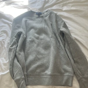 Grå sweatshirt från Polo Ralph Lauren - Klassisk grå sweatshirt från Polo Ralph Lauren med rund halsringning och broderad marinblå logga på bröstet. Tröjan har långa ärmar och ribbade muddar vid ärmslut och nederkant. Perfekt för en chill och stilren look.