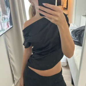 Svart oneshoulder topp i satin - Säljer en svart oneshoulder topp i glansigt satinliknande material. Toppen har en snygg draperad effekt över axeln och är croppad så magen syns. Perfekt för dig som vill ha en stilren men ändå trendig look.