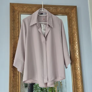 Beige skjorta från Ponje - Stilren beige skjorta från Ponje med diskreta vertikala ränder. Skjortan har bred krage, trekvartsärmar och en lös passform. Perfekt för dig som gillar minimalistisk och clean stil. Materialet känns lätt och luftigt.