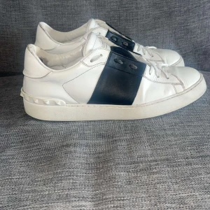 Valentino Open sneakers vit/svart skinn - Snygga Valentino Open sneakers i vitt skinn med bred svart läderdetalj över snörningen. Klassisk rund tå, platt sula och coola nitar baktill på hälen. Perfekta för dig som gillar stilrena och exklusiva sneakers med en twist.