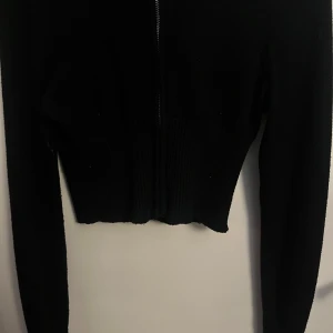 Svart croppad cardigan jacka Gina Tricot - Svart croppad cardigan jacka från Gina Tricot med huva och dragkedja framtill. Jackan är stickad med ribbad midja och långa ärmar, perfekt för en chill och stilren look.