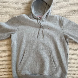 Grå hoodie från Tommy Jeans - Snygg grå hoodie från Tommy Jeans med klassisk logga broderad på bröstet. Tröjan har huva med snörning, stor magficka och ribbade muddar. Perfekt för en chill och stilren look.