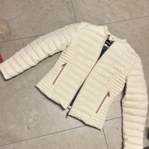 Vit pufferjacka från Rockandblue - Snygg vit pufferjacka från Rockandblue med quiltad design och dragkedjor i roséguld på fickorna. Jackan har lång ärm och en clean, modern look som passar perfekt till kalla dagar. Fodret har coolt logomönster och jackan är lätt och smidig att bära.