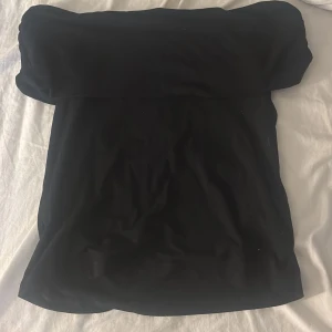 Svart axelbandslös tubtopp - Säljer en svart axelbandslös tubtopp med bandeau-modell. Toppen har en enkel och clean design med lätt rynkade kanter upptill för att sitta på plats. Perfekt att styla med jeans eller kjol för en snygg look.