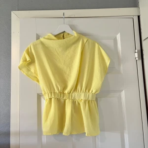 Gul blus med resårmidja och knapp från Asos - Söt ljusgul blus med korta ärmar och resår i midjan som ger en snygg volangkant. Hög halsringning med två knappar och öppning i ryggen. Luftigt material, perfekt för varma dagar.