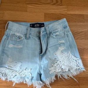 Hollister ljusblå jeansshorts med spets - Ljusblå jeansshorts från Hollister med medium hög midja slitna detaljer och råa kanter. Shortsens framsida har vit spetsdekor och snygga slitningar för en trendig look. Perfekta för sommaren och ger en chill vibe till din outfit.