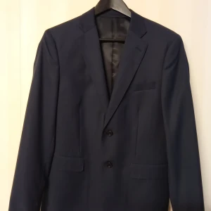 Mörkblå kavaj med Bläck märke, stl 48 - Snygg mörkblå kavaj från Bläck i modellen Hawley Blazer. Skräddad passform med två knappar framtill, klassiska fickor och helfodrad insida med vita detaljer. Perfekt för dig som vill ha en stilren och modern look.                                                                              Kan skickas med extra pris via post.                  