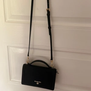 Svart axelväska från Michael Kors - Snygg svart axelväska från Michael Kors med justerbar rem och guldfärgade metalldetaljer. Väskan har ett stilrent och klassiskt utseende med handtag upptill och logga framtill. Perfekt storlek för det viktigaste och tillverkad i skinn.