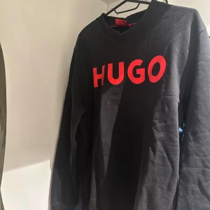 Svart tröja från Hugo med röd logga - Svart tröja från Hugo med stor röd logotyp framtill. Tröjan är lite dammig men jag kommer damma bort innan säljning. Den är i bra skick utan några skador eller liknande