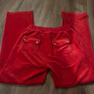 Röda velourbyxor Juicy Couture M - Röda mjukisbyxor från Juicy Couture i mjuk velour med bred resår och snörning i midjan. Byxorna har raka ben, stora fickor fram och bak med lock och silverfärgade knappar. Perfekta för en chill och trendig look.