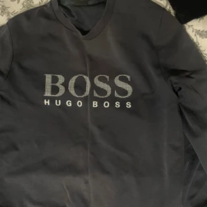Sweatshirt från Hugo Boss - Svart/väldigt mörk blå sweatshirt från Hugo Boss med stort BOSS-tryck i vitt framtill. Klassisk rund halsringning och långärmad modell. Tröjan är lite skadad vid ena armbågen, men det är inte så synligt (se bild ovan) 