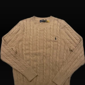Beige kabelstickad tröja Polo Ralph Lauren - Kabelstickad beige tröja från Polo Ralph Lauren med klassisk rund halsringning och broderad marinblå logga på bröstet. Tröjan är långärmad och har ribbade muddar vid ärmslut och nederkant. Perfekt för lager-på-lager under kyliga dagar. Använt en gång. Nypris: 2400kr 
