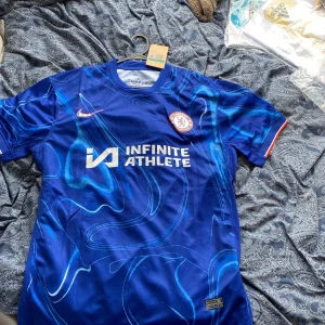 Chelsea FC blå matchtröja Enzo #8 XL - Chelsea FC:s officiella matchtröja i blått med unikt abstrakt mönster, tryck 'INFINITE ATHLETE' på bröstet och Enzo #8 på ryggen. Nike Dri-FIT-material för bästa komfort. Chelsea-märke på bröstet och röda detaljer på ärmsluten. Tröjan är helt i nyskick och har även lappar kvar. Storleken är XL och passar någon som är 180-185 cm lång. Vid minsta fundering hör av dig.