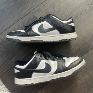 Nike Dunk Low svart/vit EUR 44 - Säljer ett par Nike Dunk Low i klassisk svart och vit färg. Skorna har svart läder på ovandelen, vita paneler och svart swoosh. Lågt skaft, svart snörning och vit mellansula. Perfekta för dig som gillar streetwear och vill ha en ikonisk sneaker i garderoben.