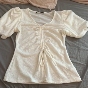 Vit blus med puffärm från Bikbok - Supersöt vit blus från Bikbok med puffiga korta ärmar och v-ringning. Toppen har snygg snörning framtill som ger en rynkad effekt och markerar midjan. Materialet är mjukt och har en lätt struktur som gör den extra intressant. Perfekt för dig som gillar romantiska detaljer!