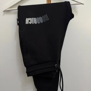 Svarta mjukisbyxor från Hoodrich - Svarta mjukisbyxor från Hoodrich med broderad logga i vitt och grått på ena benet. Byxorna har dragsko i midjan och sidofickor, perfekta för en chill och avslappnad stil.