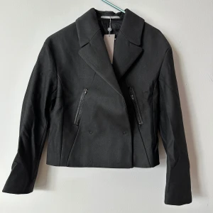 Svart kavajjacka med dragkedja från Tiger of Sweden  - Stilren svart kavajjacka med breda slag, croppad passform och coola dragkedjor både fram och bak samt på ärmarna. Jackan har två sneda fickor med dragkedja och en clean look som passar till många outfits. Perfekt för dig som gillar en edgy men klassisk stil. Passar en 34-36. Nypris 5500kr 