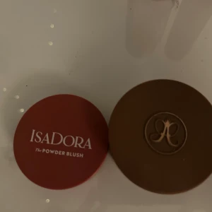 Isadora rouge & Anastasia bronzer - Säljer ett puderrouge från Isadora i en snygg mörkrosa nyans och en bronzer från Anastasia Beverly Hills i en varm brun ton. Båda kommer i runda dosor, rouget i röd plast och bronzern i brun dosa med guldig logga. Perfekt för att skapa glow och färg i ansiktet. Säljer för har dubbla av båda. (Säljer för VÄLDIGT bra pris !!❤️‍🔥❤️‍🔥