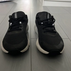 Svarta Under Armour löparskor strl 44 - Snygga svarta löparskor från Under Armour med vit logga på sidan och vit/grå sula. Ovandelen är i mest för bra andningsförmåga och skorna har klassisk snörning. Perfekta för träning eller en sportig vardagsstil.