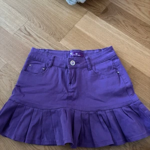 Lila volangkjol från Red Rose - Superfin lila kortkjol från Red Rose med volangkant nertill och klassiska jeansfickor både fram och bak. Kjolen har bälteshällor, knapp och dragkedja framtill samt snygga detaljer på bakfickorna. Perfekt för dig som vill sticka ut lite extra!